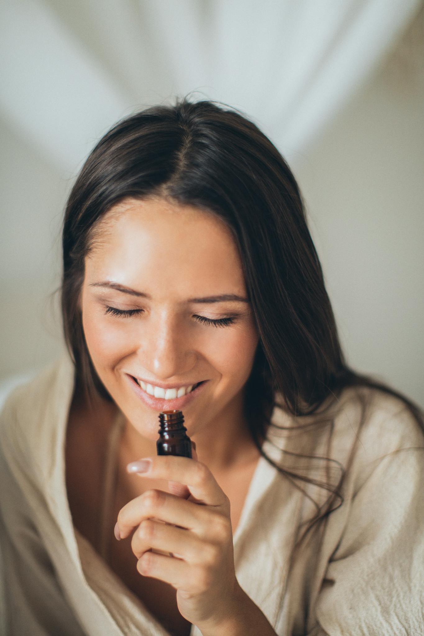 A smiling woman with closed eyes enjoying the scent from a glass bottle, highlighting a moment of relaxation and aroma therapy.